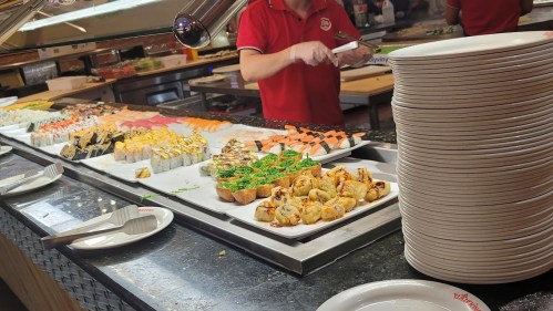 A buffet display featuring various sushi and rolls, with a server in a red shirt and stacks of empty plates nearby.