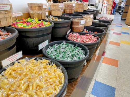 A colorful display of various candies in large black containers on a wooden table, with a tiled floor in the background.
