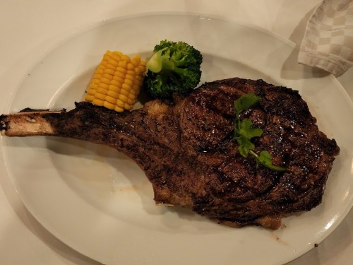 A large, grilled ribeye steak with a side of corn and broccoli on a white plate.