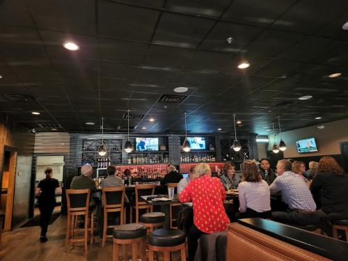 A busy restaurant bar with patrons seated at tables, a bartender, and multiple TVs showing sports.