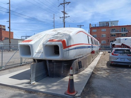 A weathered, futuristic train-like vehicle sits on a concrete platform, surrounded by a fence and parked cars.