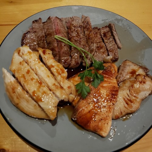 A plate of grilled meats featuring slices of steak, chicken, and salmon, garnished with a sprig of cilantro.