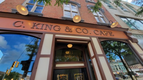 Sign for C. King & Co. Café on a brick building, with large windows reflecting the sky and surrounding trees.