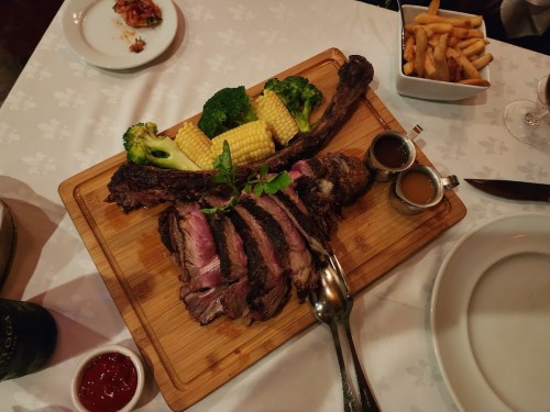 A wooden platter with sliced steak, broccoli, corn, dipping sauces, and a side of fries on a table.
