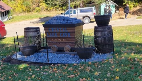 A decorative mining cart with blue stones, surrounded by barrels and plants, set in a grassy area with trees in the background.