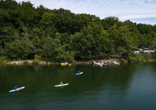 Four people paddle on a calm lake surrounded by lush green trees and a small dock in the background.