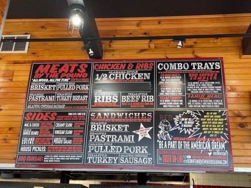 Menu board featuring various meat options, sides, sandwiches, and combo trays in a rustic restaurant setting.