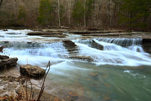 A serene river flows over layered rocks, creating gentle waterfalls surrounded by trees in a tranquil forest setting.