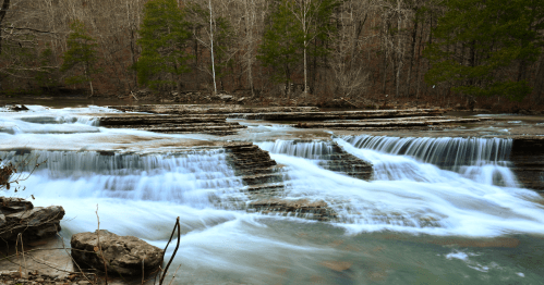 A serene waterfall cascades over rocky ledges, surrounded by trees in a tranquil forest setting.