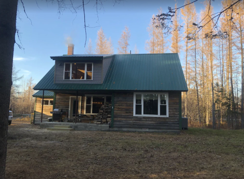 A rustic two-story cabin with a green metal roof, surrounded by trees and a clear blue sky.