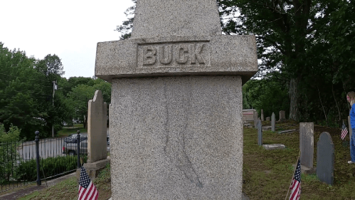 A granite gravestone with the name "BUCK" engraved, surrounded by a cemetery and a small American flag.