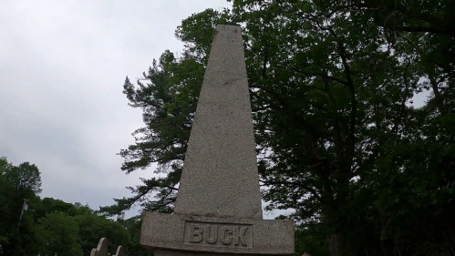 A tall, stone obelisk monument with the name "BUCK" engraved at its base, surrounded by trees under a cloudy sky.