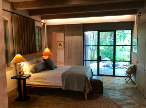 Cozy bedroom with a bed, lamps, and large windows overlooking a green outdoor space. Natural light fills the room.