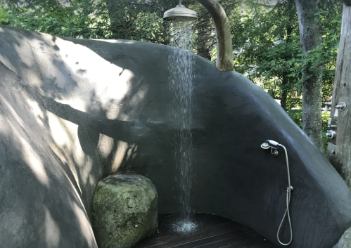 Outdoor shower with a unique stone wall, water cascading from a round showerhead, surrounded by greenery.