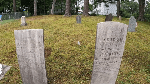 Two weathered gravestones in a grassy cemetery, surrounded by trees, with inscriptions partially visible.