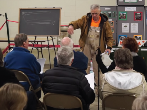 A speaker in a brown jacket gestures while presenting to an audience seated in folding chairs. A chalkboard is visible.