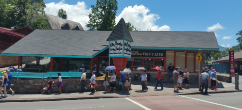 A colorful candy shop with a peaked roof, surrounded by people walking on a sunny day. Trees and blue sky in the background.