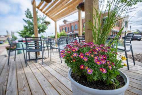 A vibrant flower pot with pink and yellow blooms on a wooden patio, with empty chairs and a building in the background.