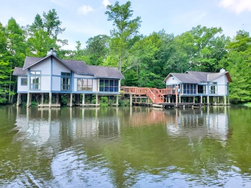 Two houses on stilts by a calm lake, surrounded by lush green trees and a wooden walkway connecting them.