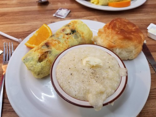 A plate with an omelet, a biscuit, and a bowl of grits topped with butter, alongside a slice of orange.