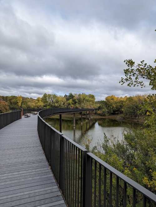 A winding wooden walkway curves over a calm river, surrounded by trees with autumn foliage under a cloudy sky.