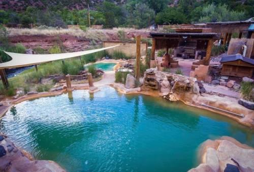 A serene outdoor hot spring pool surrounded by natural rock formations and greenery, with a shaded area above.