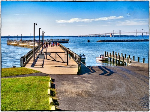 A scenic view of a pier extending into the water, with boats and a bridge in the background under a clear blue sky.