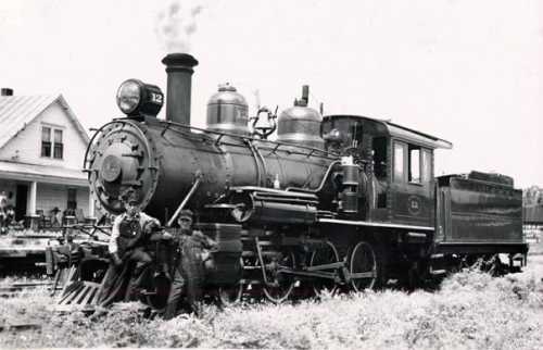 A vintage steam locomotive with two men posing beside it, set against a backdrop of a house and greenery.