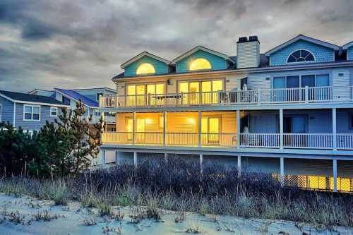 A beach house at dusk, with glowing windows and a sandy foreground, surrounded by cloudy skies.