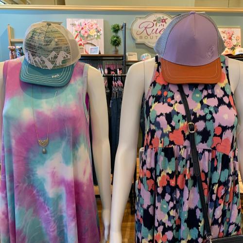 Two colorful dresses on display, one tie-dye and one patterned, paired with matching hats in a boutique setting.