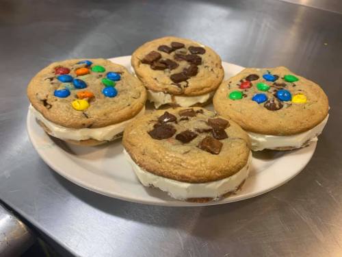 Four cookie sandwiches with colorful candy and chocolate chunks, filled with creamy frosting, on a white plate.
