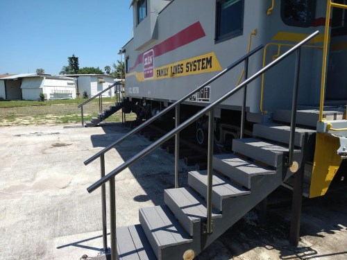 A train car with a yellow and red logo, featuring metal stairs leading up to it, set in a grassy area.