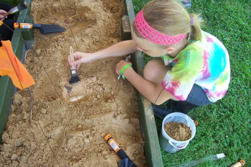 A child in a tie-dye shirt brushes sand while digging in a garden bed, surrounded by tools and a bucket.