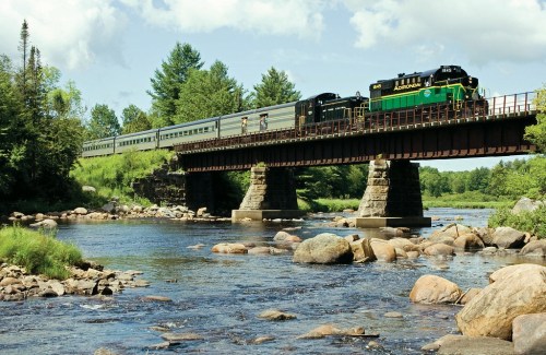 A train crosses a bridge over a river, surrounded by trees and rocky banks under a partly cloudy sky.
