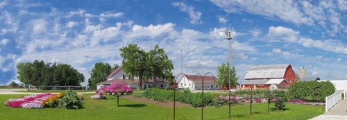 A panoramic view of a farm with colorful flower beds, a red barn, and a windmill under a blue sky with fluffy clouds.