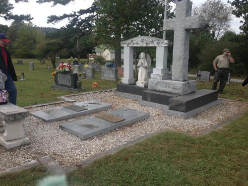 A cemetery scene featuring a gravestone with a cross, a statue, and flowers, surrounded by grass and trees.
