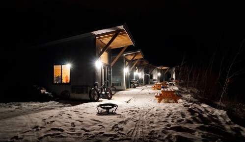 Cozy cabins illuminated at night, surrounded by snow, with picnic tables and bicycles nearby.