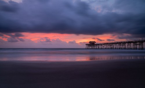 A serene beach scene at sunset, featuring a pier silhouetted against colorful clouds and calm ocean waves.