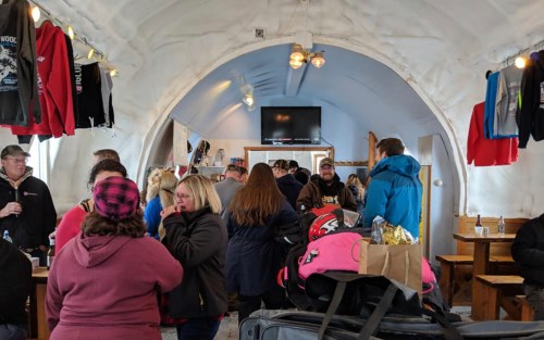 A busy indoor space with people socializing, wearing winter clothing, and various items on tables.