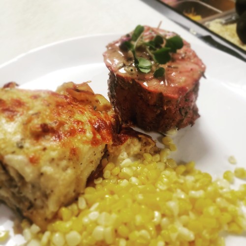 A plate featuring creamy lasagna, a meatloaf slice topped with sauce, and a side of yellow corn.