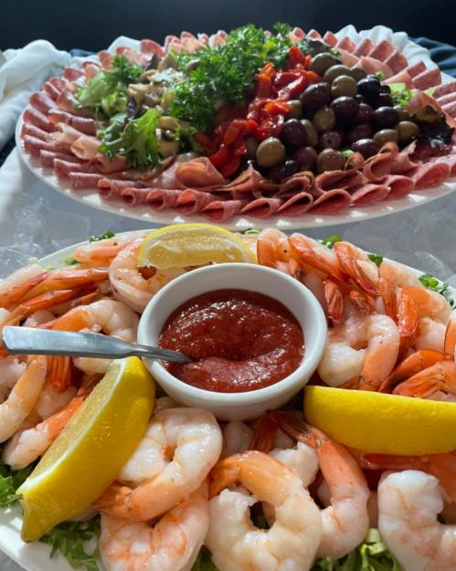 A platter of shrimp with cocktail sauce and lemon, alongside a meat and olive salad platter.