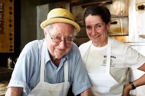 A smiling older man in a straw hat and a woman in a white shirt and apron stand together in a kitchen.