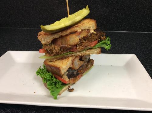 A tall sandwich stacked with layers of lettuce, tomato, and crispy ingredients, topped with a pickle slice.