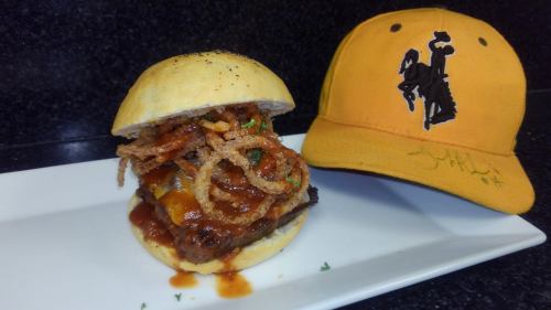 A gourmet burger topped with crispy onions and sauce, next to a yellow cap featuring a cowboy logo.