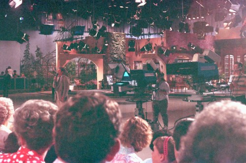 A television studio set with cameras and crew, featuring a colorful backdrop and an audience in the foreground.