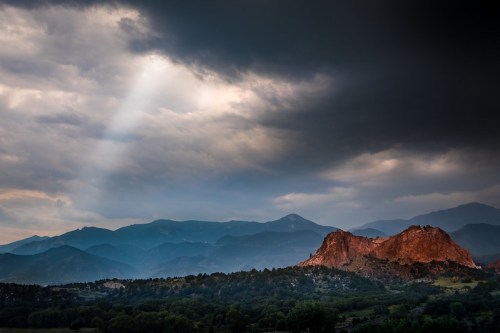 A dramatic landscape featuring red rock formations under a cloudy sky with a beam of light breaking through.