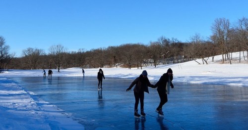 People ice skating on a frozen lake surrounded by snow-covered trees under a clear blue sky.