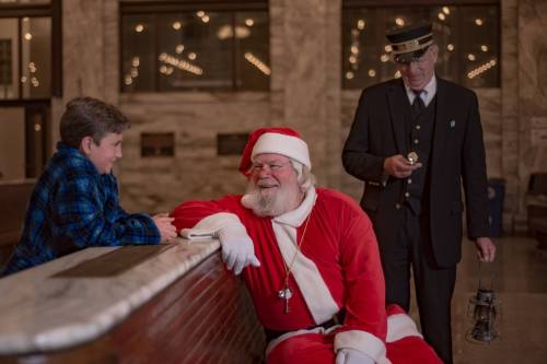 A cheerful Santa Claus sits on a bench talking to a young boy, while a train conductor stands nearby holding a bell.