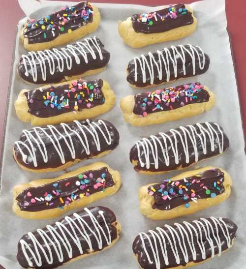 A tray of eclairs topped with chocolate icing, white drizzle, and colorful sprinkles on parchment paper.