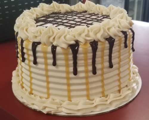 A round cake with white frosting, topped with chocolate and caramel drizzles, and decorative swirls around the edges.